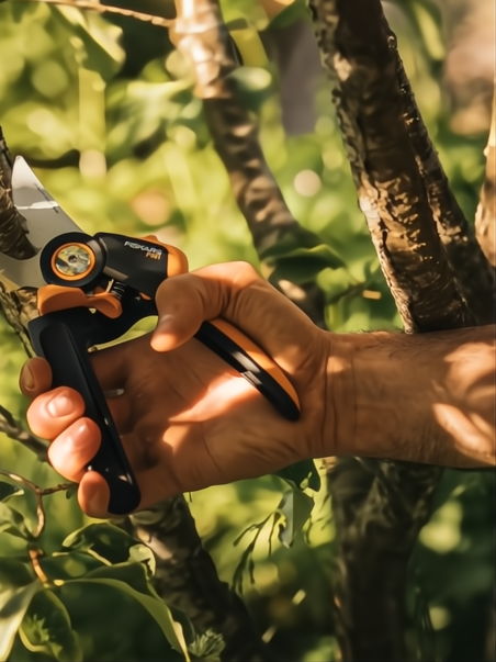 Mano sosteniendo tijera de podar Fiskars X-Series P961 negra y naranja, con hoja de acero, podando una rama.