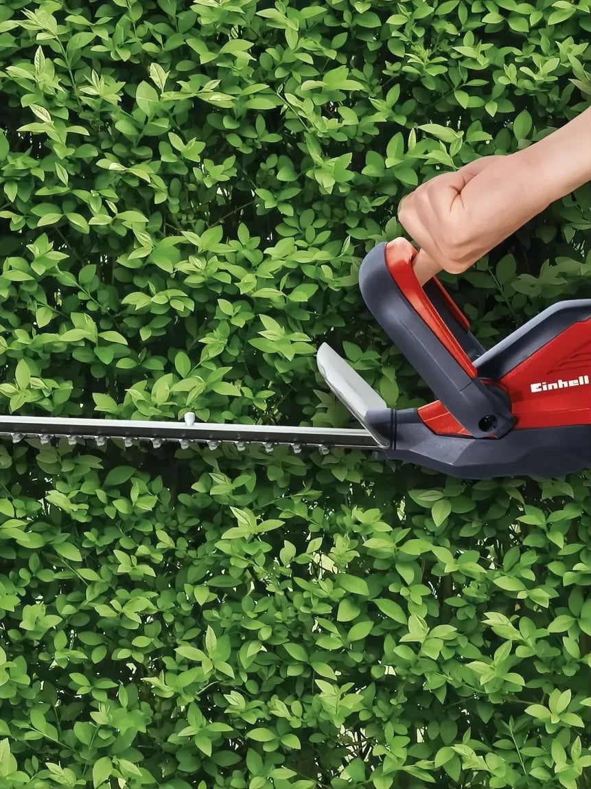 Red and black Einhell cordless hedge trimmer cutting a green hedge.