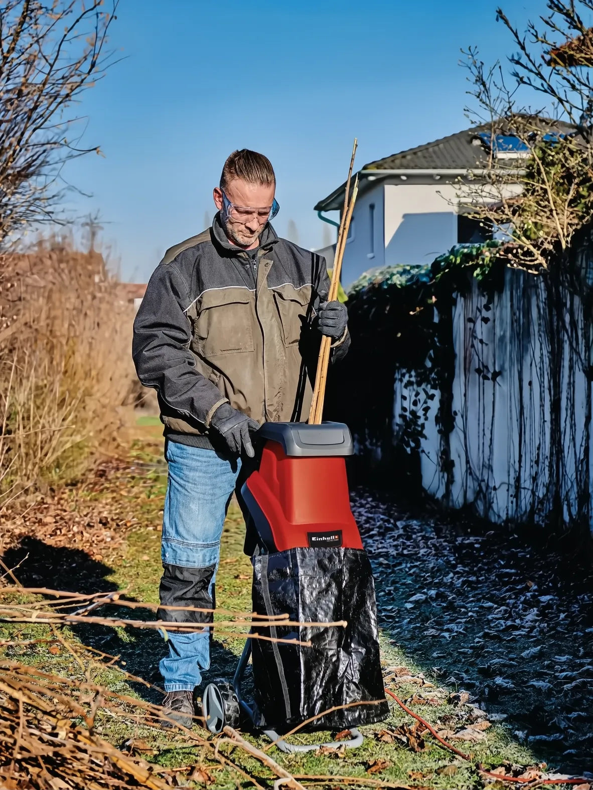Hombre usando triturador eléctrico Einhell GC-KS 2540 rojo y negro con bolsa de residuos en jardín.